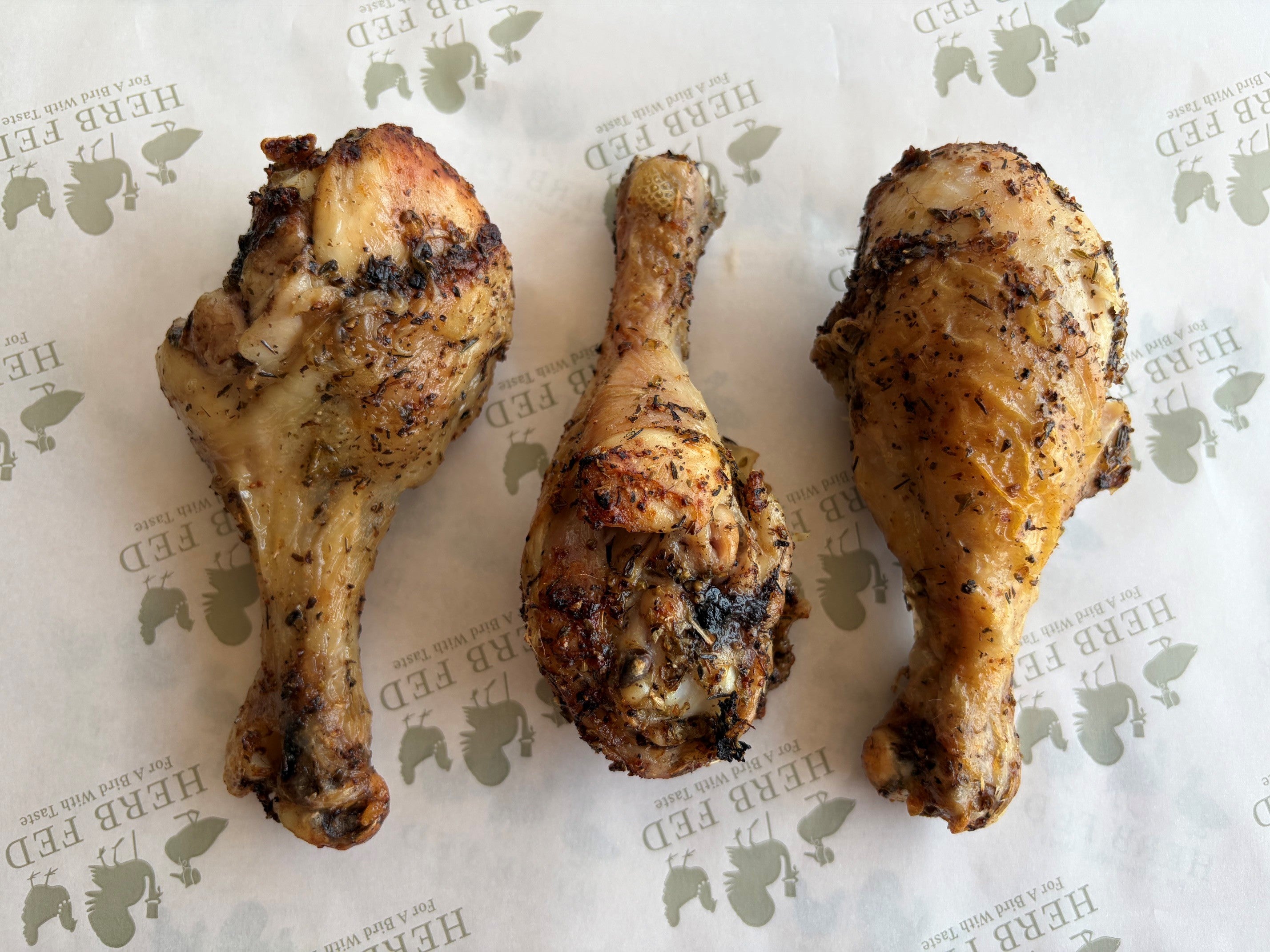 Three roasted Herb Fed chicken drumsticks with charred, crispy skin are arranged side-by-side.