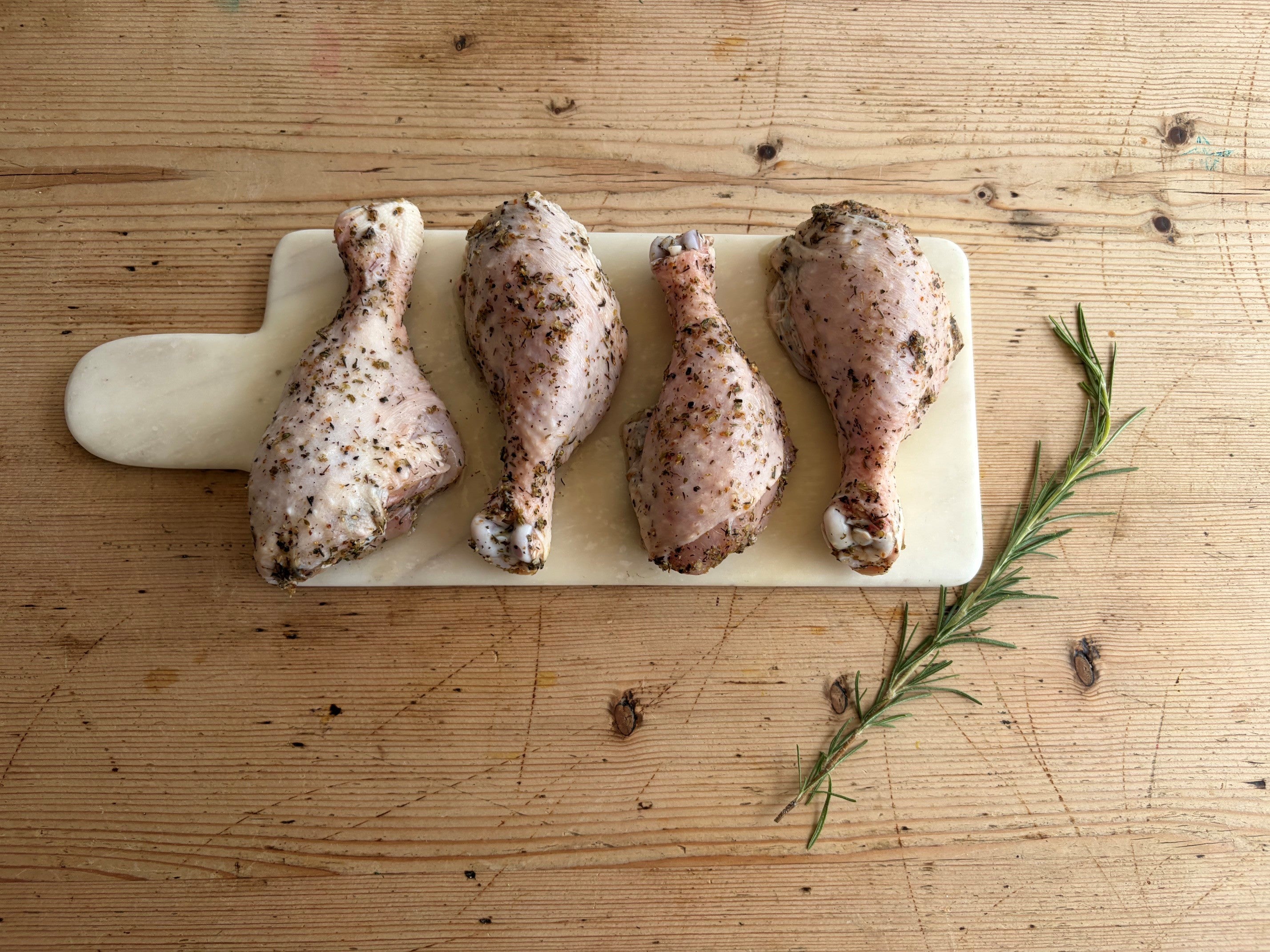 Four raw Herb Fed chicken drumsticks seasoned with Gyros herbs and spices, arranged on a rectangular white cutting board over a wooden table, with a rosemary sprig to the right.