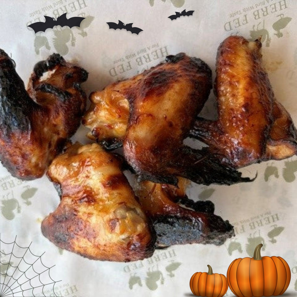 Four glazed chicken wings with charred edges, arranged on patterned Herb Fed paper with Halloween-themed graphics including bats, a spiderweb, and pumpkins. Perfect for a Halloween feast.
