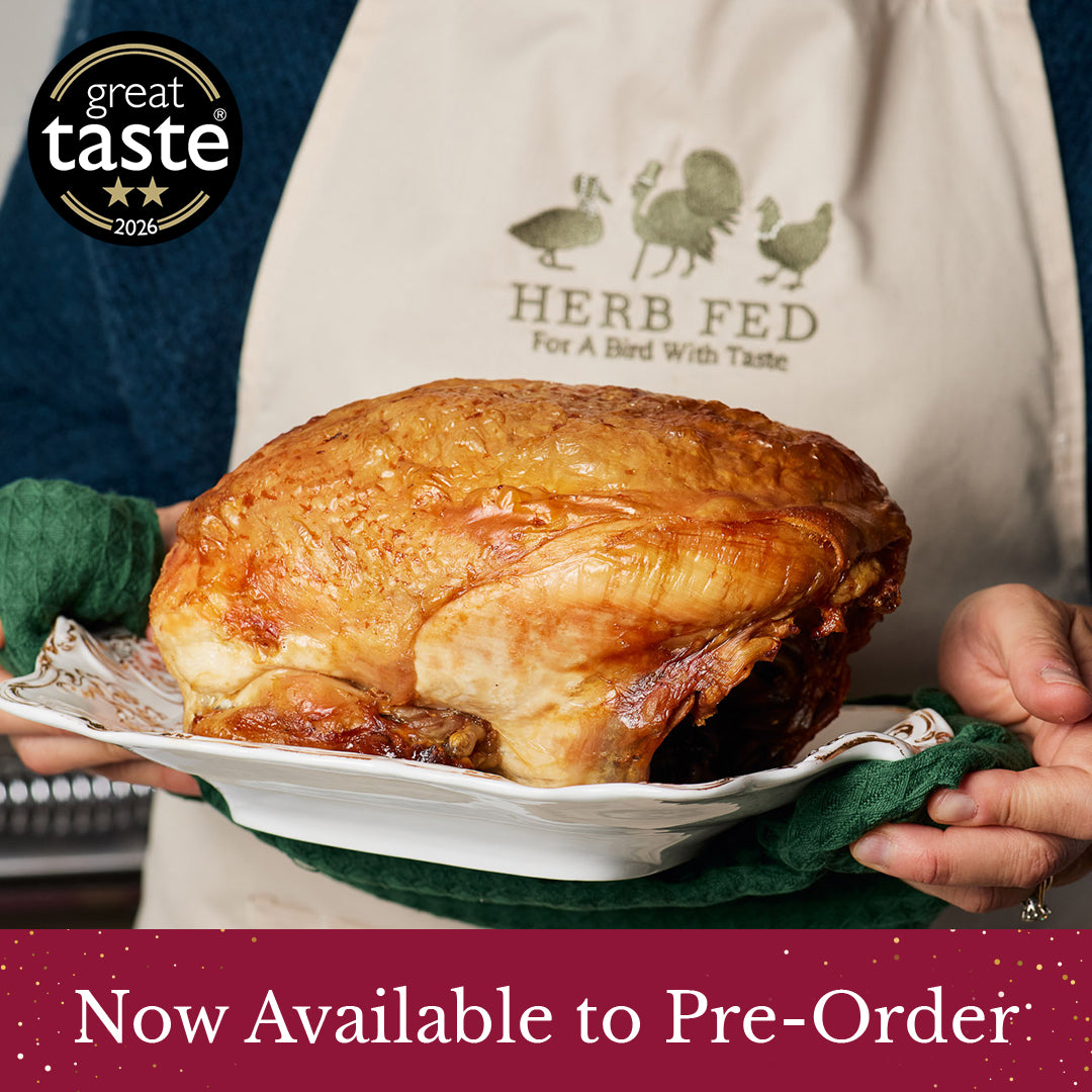 A roasted Christmas Rooster Crown on a white platter held by hands wearing a cream apron with 'HERB FED' printed on it. A '2 star great taste 2026' badge appears in the top-left corner, and a maroon banner at the bottom reads 'Now Available to Pre-Order'.