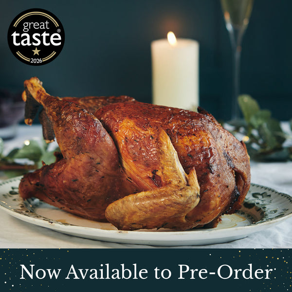 A roasted whole Herb Fed bronze turkey on a white plate in a dinner setting with a lit candle, glass, and greenery in the background. A black 'great taste 2026' badge appears in the top-left, and a dark banner at the bottom reads 'Now Available to Pre-Order.