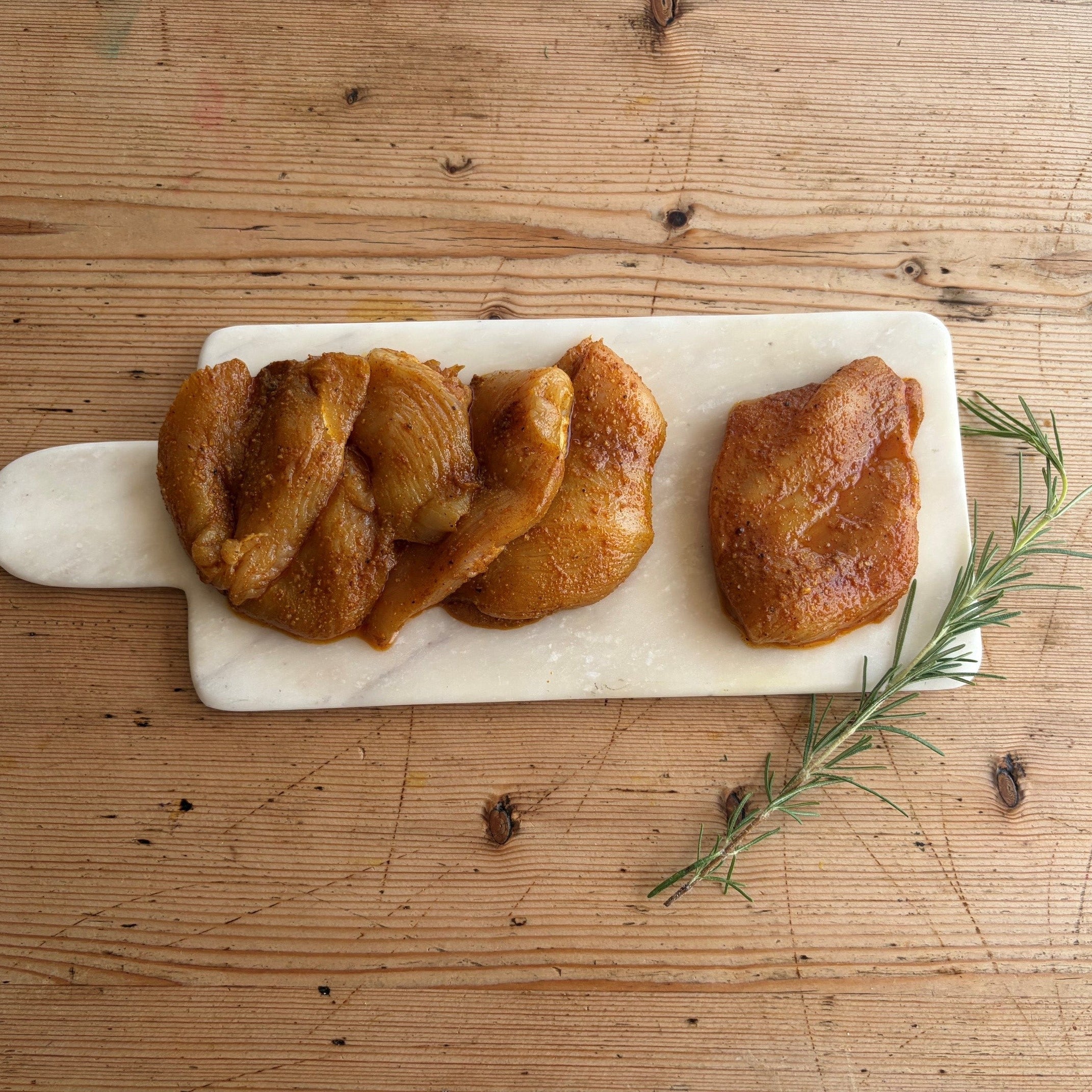 Three pieces of glazed roasted chicken breast arranged on a white marble serving board with a handle, placed on a rustic wooden table with a rosemary sprig nearby.