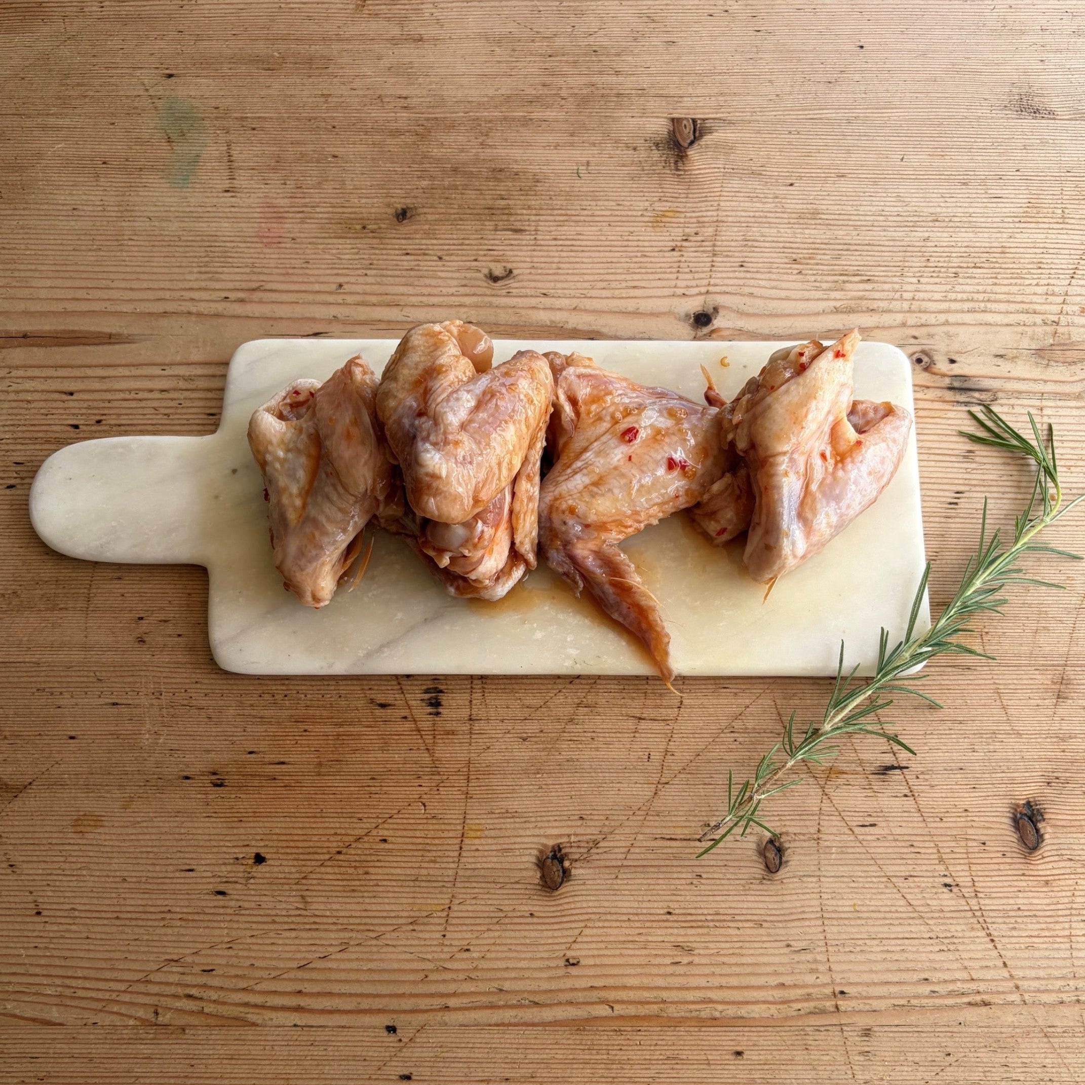 Raw marinated Herb Fed chicken wings arranged on a small white cutting board on a rustic wooden table, with a sprig of rosemary placed beside the board.