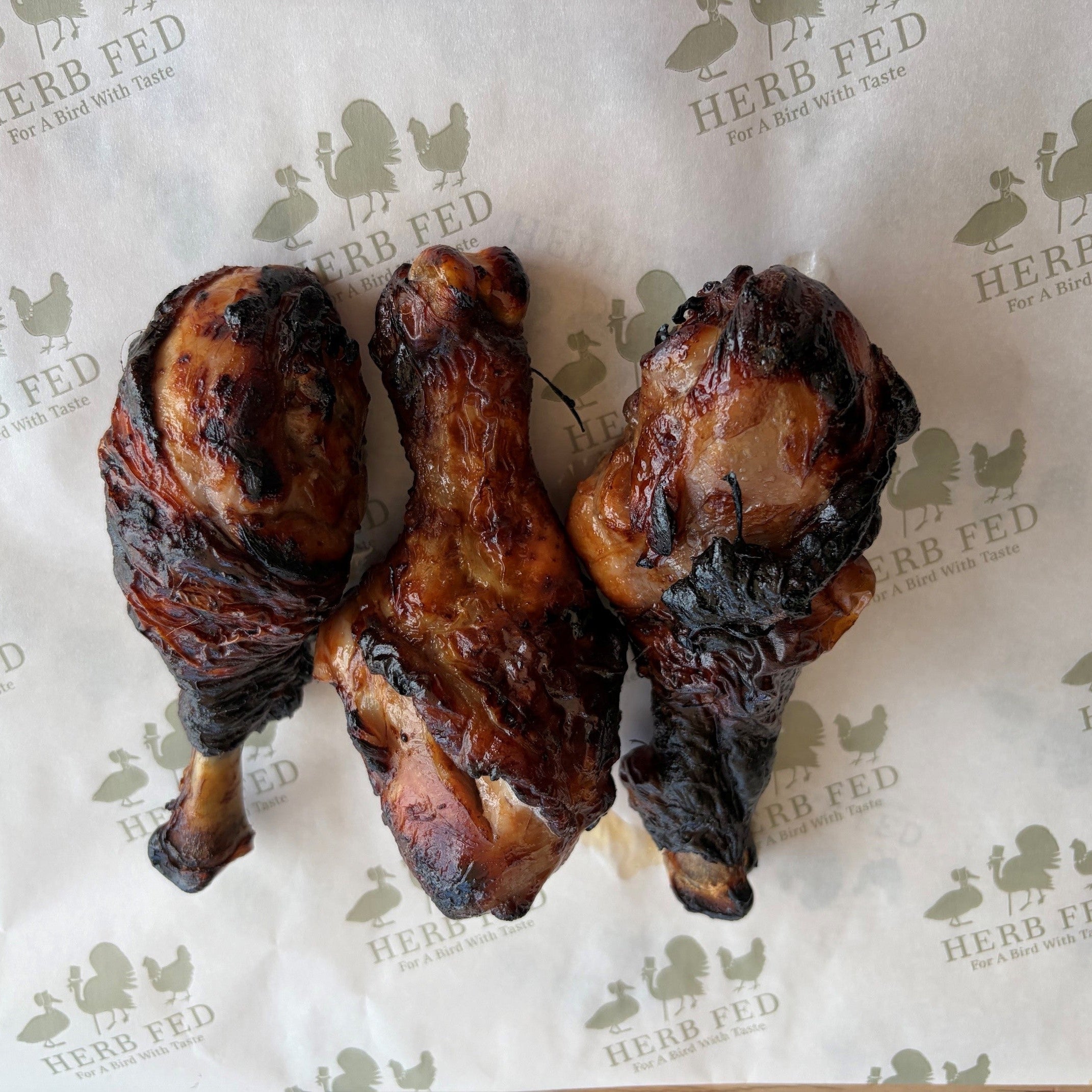 Three grilled Herb Fed chicken drumsticks charred to perfection arranged on a table.