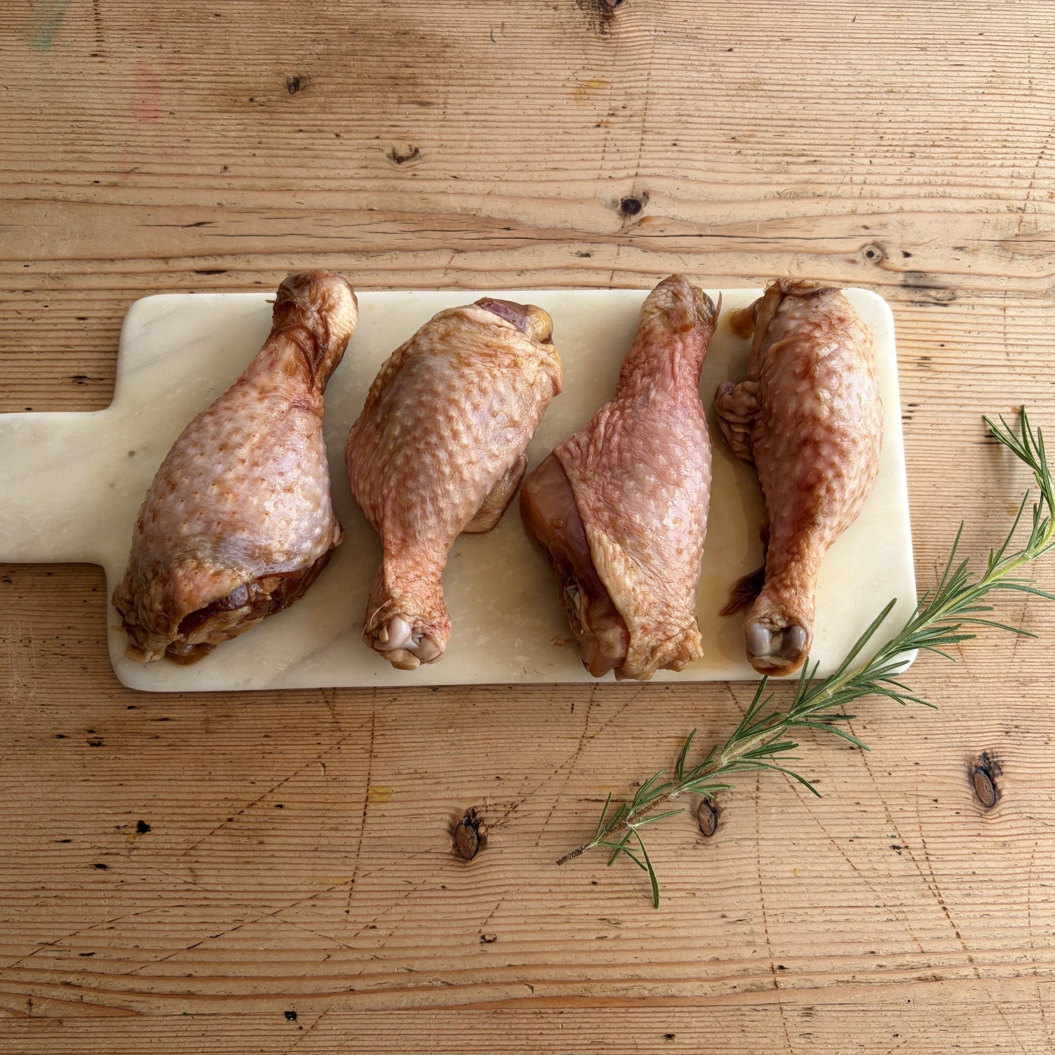 Four raw chicken drumsticks arranged vertically on a light beige cutting board, placed on a rustic wooden table with a rosemary sprig nearby.