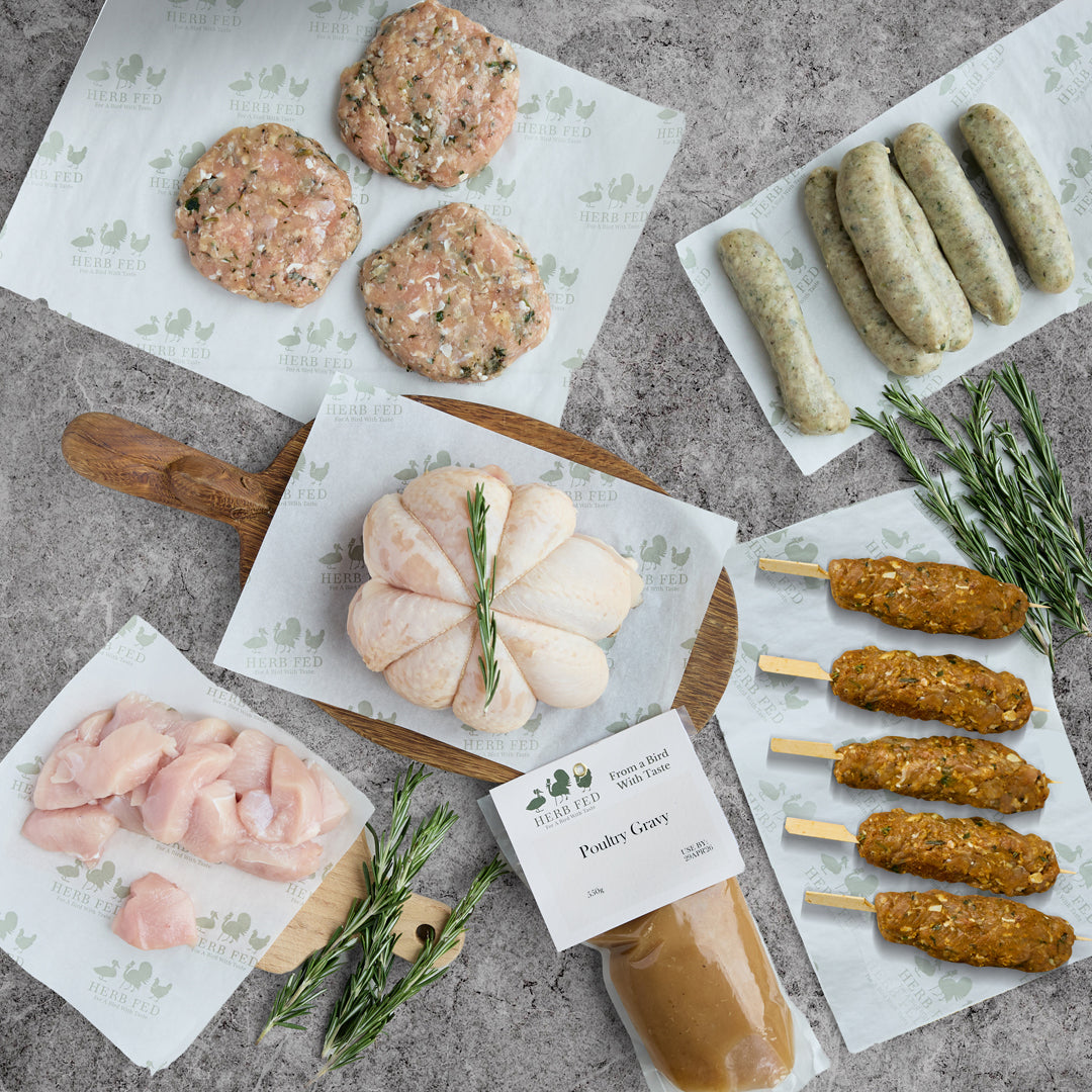 An image of the Herb Fed Winner Winner Chicken dinner box contents showing various raw ingredients on white paper sheets with green branding. The contents includes gravy, chicken burgers, chicken sausages, diced chicken breasts, stuffed chicken cushion, chicken shawarma's all arranged on a grey textured surface.