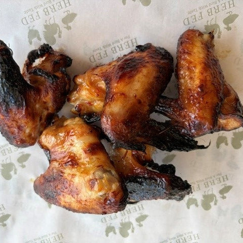 Four grilled chicken wings with charred edges and a glossy glaze, arranged on branded Herb Fed paper.